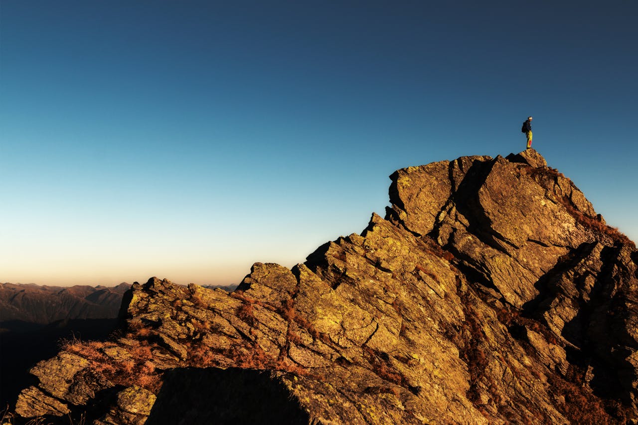 our-services-1 A lone hiker stands triumphantly atop a rocky mountain peak at sunrise, embracing nature and the view.